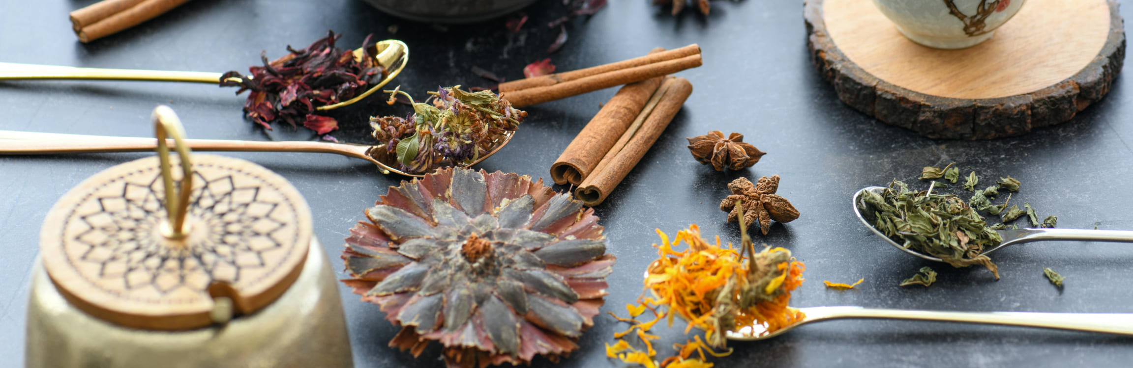 Tea brewing scene with a black teapot, floral ceramic cup, and spoons of herbs and spices like cinnamon, star anise, and hibiscus on a dark surface.