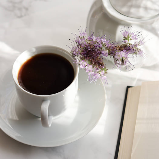 A white cup of black coffee sits on a saucer next to light purple flowers and an open book. The scene is calm and inviting, with soft lighting.