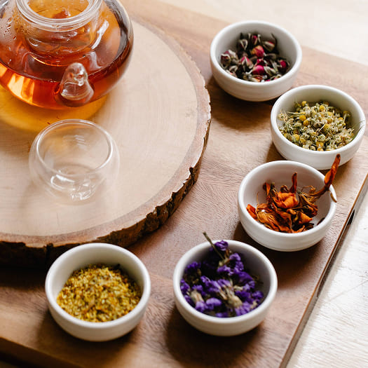 A wooden tray holds a glass teapot with herbal tea and five small bowls containing dried flowers and herbs, creating a warm, natural, and inviting scene.