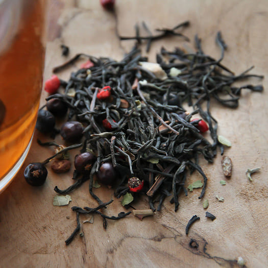 A close-up of loose leaf tea with herbs and berries on a wooden surface. A glass of brewed tea is partially visible, creating a warm and inviting feel.