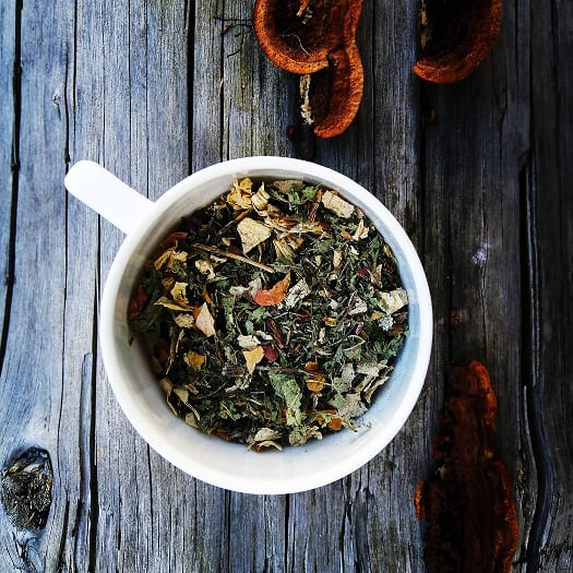 A white mug filled with assorted dried tea leaves sits on a rustic wooden table, accompanied by scattered pieces of dried fungi, evoking a natural, earthy tone.