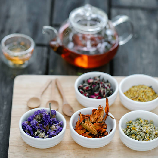 A wooden board with bowls of dried herbal tea blends, including purple and orange flowers. A glass teapot with brewed tea is in the background, creating a calm, niche tea scene.