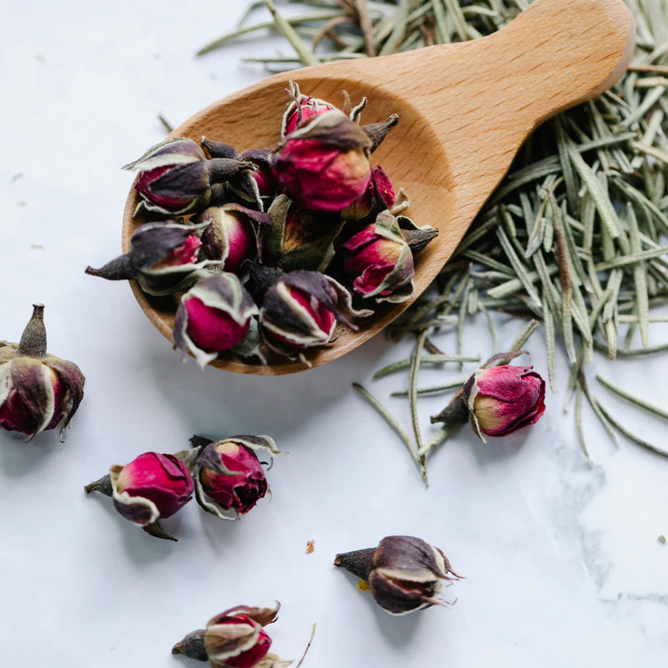 Wooden scoop with dried rosebuds and rosemary sprigs on a white surface, creating a natural, aromatic, and rustic aesthetic.