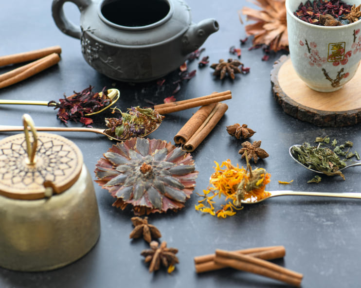 Tea brewing scene with a black teapot, floral ceramic cup, and spoons of herbs and spices like cinnamon, star anise, and hibiscus on a dark surface.
