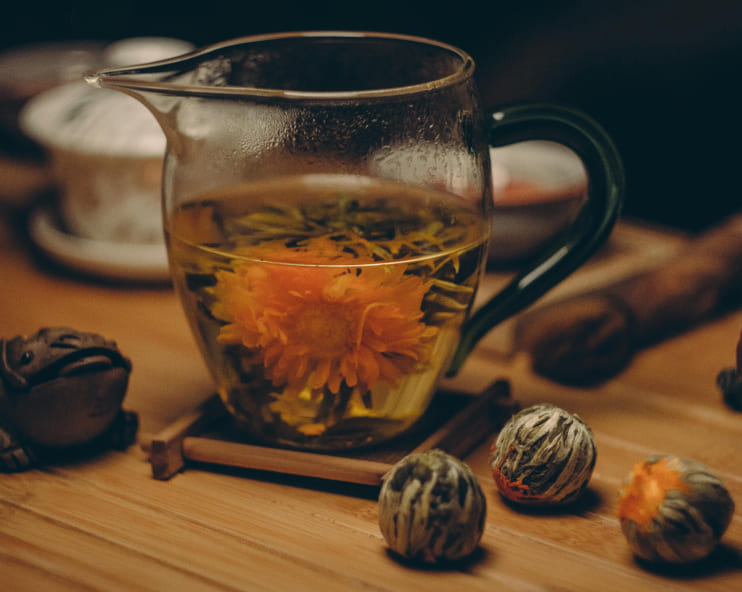 A glass pitcher with blooming tea, featuring a vibrant orange flower, sits on a wooden tray. The setting has a warm, cozy ambiance.