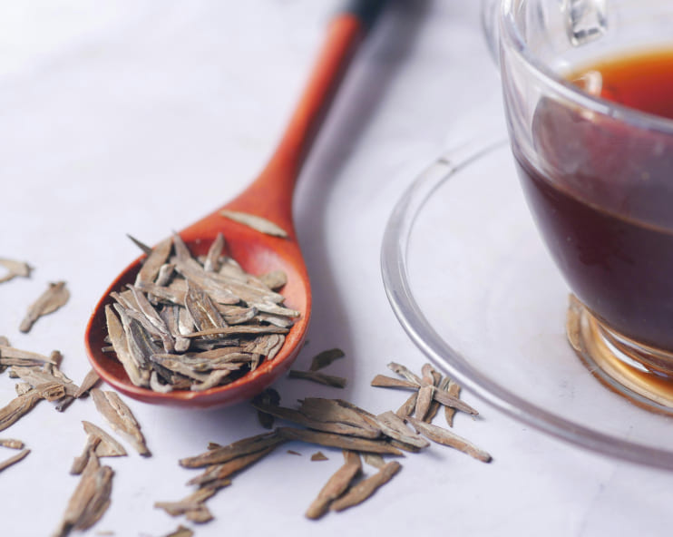 A wooden spoon with dried tea leaves next to a clear glass cup filled with dark tea. The scene conveys a warm, calming tea preparation ambiance.