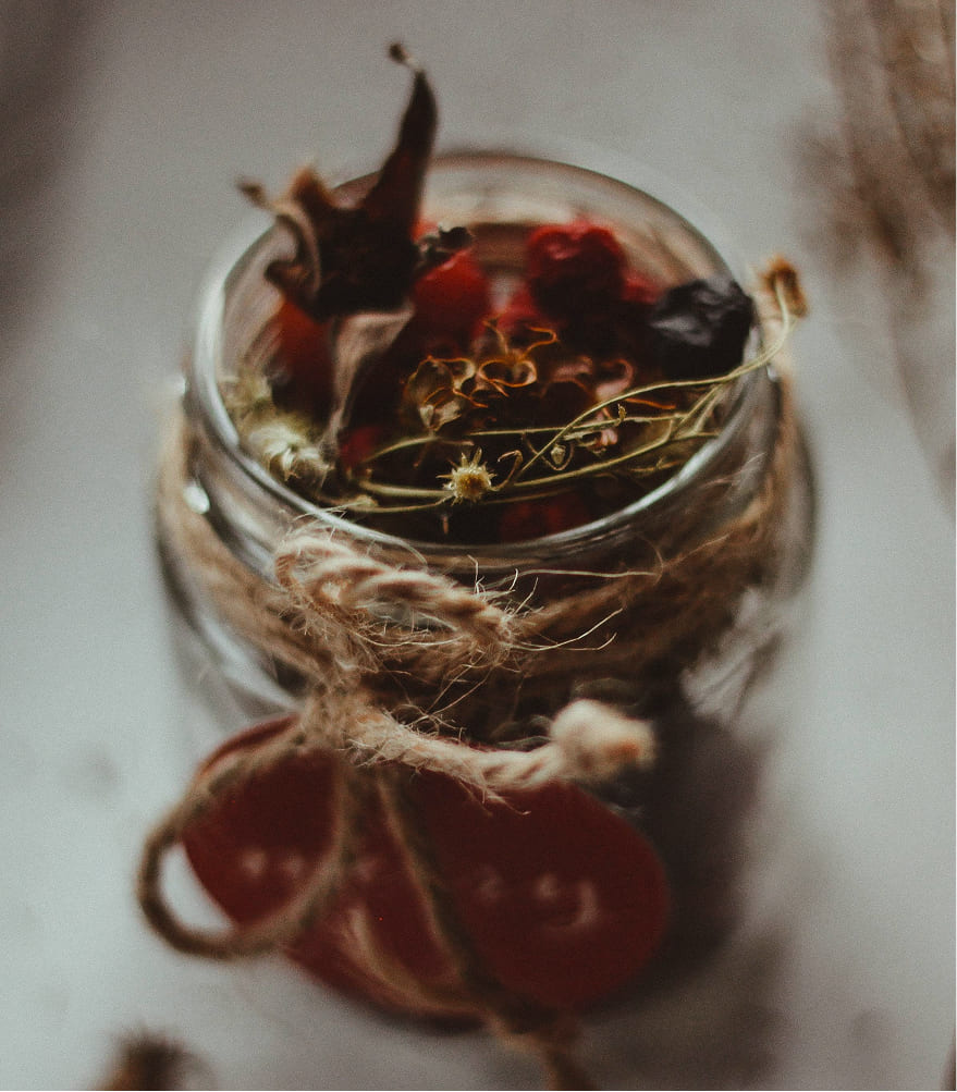 A rustic glass jar filled with dried herbs and flowers, wrapped with a twine bow. The scene gives a warm, vintage feel on a soft-focus background.