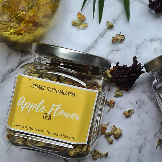 Labeled jar of 'Apple Flower Tea' by Organic Touch Malaysia on marble. Dried flowers and a glass teapot with brewed tea are beside the jar.
