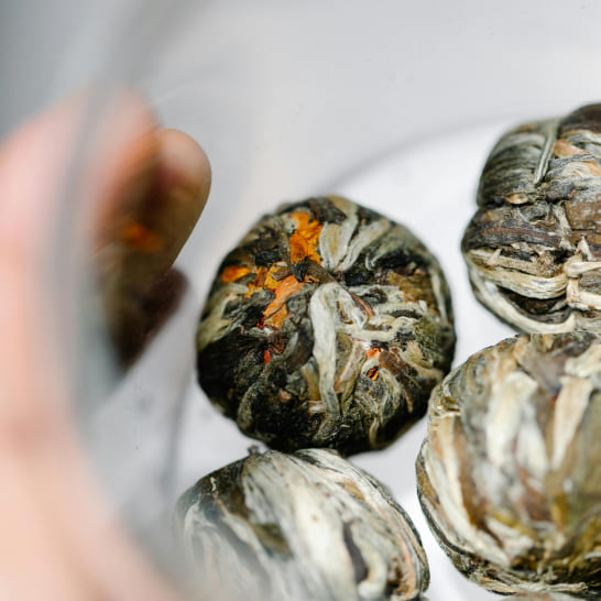 Close-up of dried flower tea balls with intertwined leaves and petals in shades of green, white, and orange, creating an intricate, natural texture.