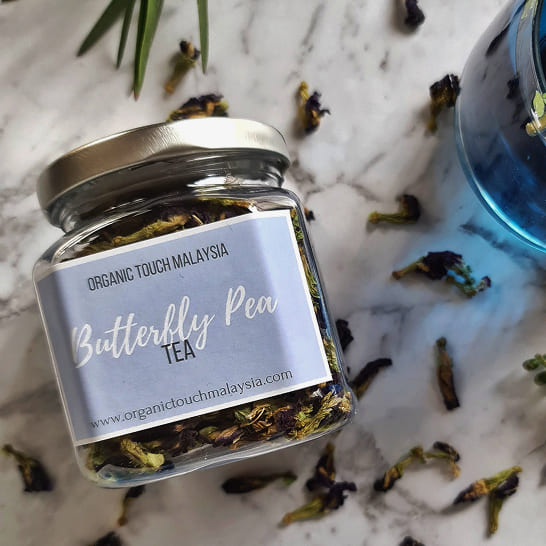 A jar of 'Butterfly Pea Tea' on a marble surface, with scattered dried flowers and a vibrant blue liquid nearby. The scene is fresh and inviting.