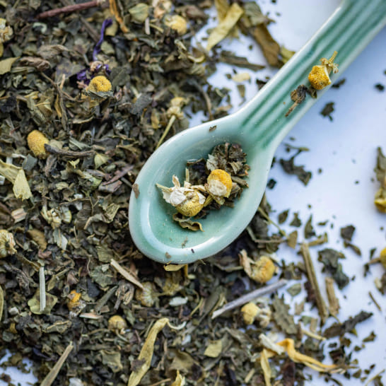 A light green ceramic spoon rests on a bed of dried chamomile and herbal tea leaves, creating a soothing, natural atmosphere.