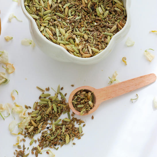 A white bowl filled with cumin sits on a light surface. A wooden scoop and scattered seeds create a rustic, aromatic scene.