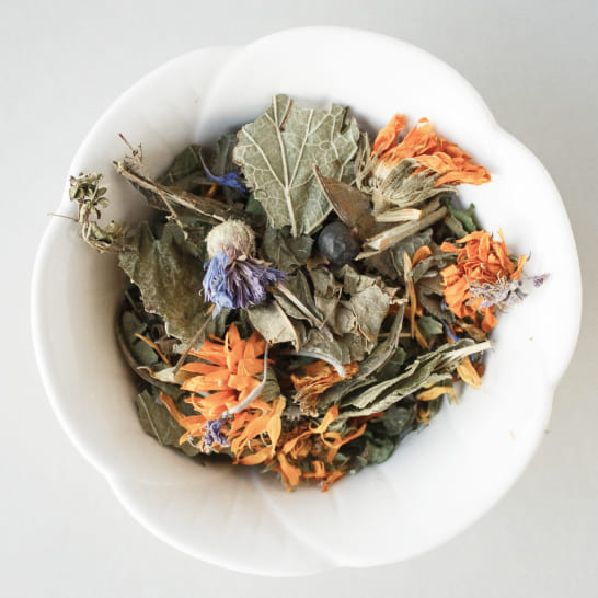 A white bowl filled with a mix of dried herbs and flowers, including green leaves and vibrant orange and purple petals, on a neutral background.
