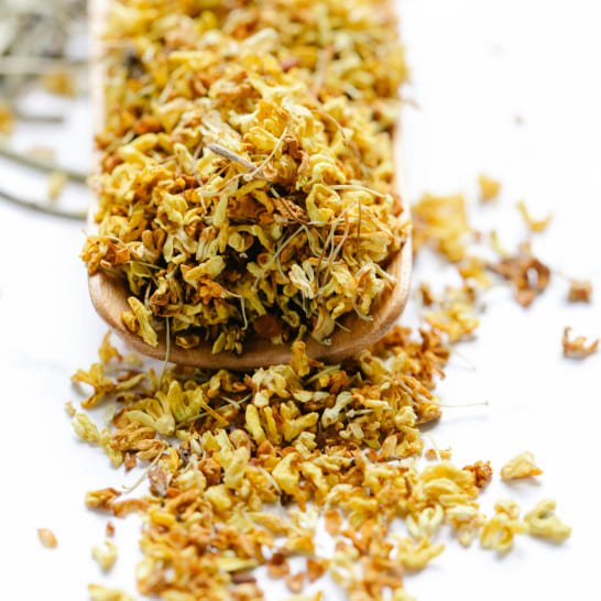 A wooden scoop overflows with dried yellow flowers against a white background, creating a warm, earthy texture that evokes a sense of calm.