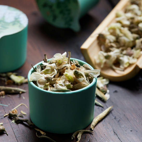 A teal container filled with dried jasmine flowers sits on a dark wooden table. Nearby, more flowers spill from a wooden scoop, creating a rustic, aromatic scene.