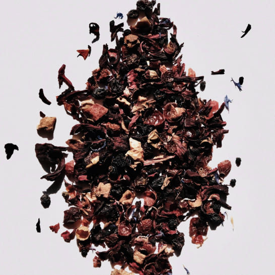 A pile of loose-leaf dried herbal tea blend scattered on a white surface. The tea consists of various dried fruits and leaves in shades of red and brown.
