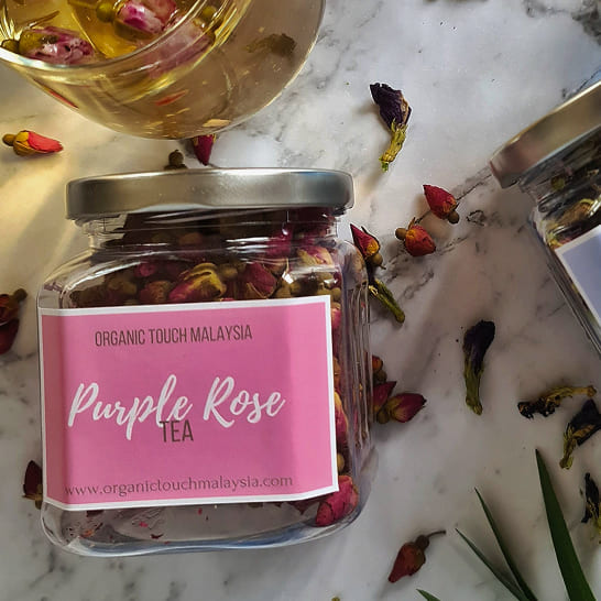 Jar of 'Purple Rose Tea' by Organic Touch Malaysia on marble, surrounded by dried rosebuds. A glass of brewed tea adds a warm, inviting glow.