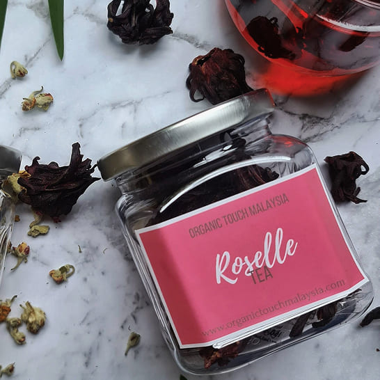 Jar of Roselle tea on a marble surface, surrounded by dried petals and a glass of red tea. The scene conveys freshness and natural essence.