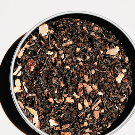 A round tin filled with loose black tea leaves mixed with dried orange peel, cinnamon bits, and spices. The lighting creates a warm, inviting tone.