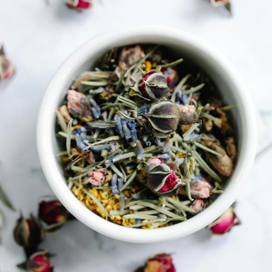 A white bowl filled with a colorful herbal tea blend, including dried rosebuds, lavender, and green leaves. Scattered rosebuds surround the bowl.
