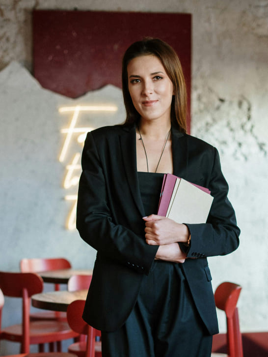 Young woman in a black suit holds books, standing confidently in a stylish cafe with red chairs and a neon sign in the background, conveying a professional tone.