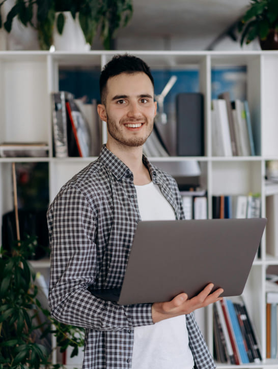 Smiling person holding a laptop, stands in a room with white bookshelves filled with books and decor. Plants add a touch of greenery, conveying a cozy and productive atmosphere.