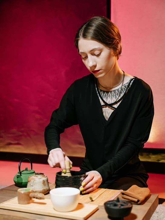 A person in a black outfit ceremonially prepares matcha tea at a table. A green teapot, a ceramic cup, and other tea utensils are also present. The background glows warmly with red and pink hues, creating a serene atmosphere.
