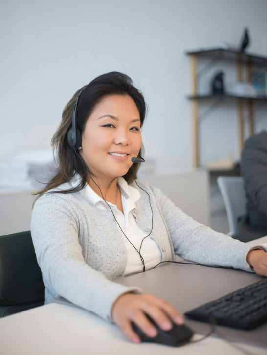 Smiling woman with a headset sits at a desk, using a computer mouse. The office setting appears professional and organized, conveying efficiency.