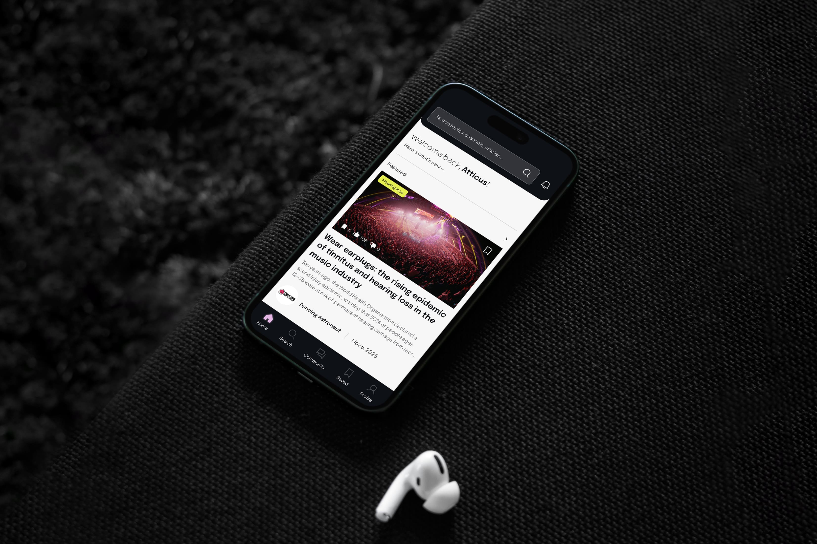 Smartphone on a black couch displays an article card with concert image. White wireless earbuds lie nearby, creating a sleek, modern feel.